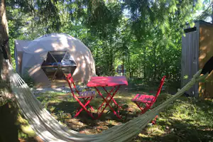 Glamping-Zelt im Wald mit rotem Tisch und Stühlen, Hängematte im Vordergrund und Holzgebäude am Rand.