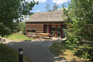 Holzhütte mit Terrasse, Holztisch und Bänken, umgeben von Bäumen und Grün am Weg auf dem Campingplatz.