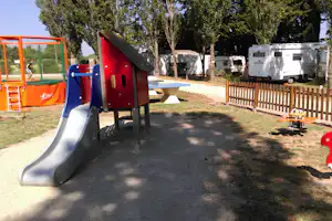 Spielplatz mit Rutsche und Kletterelementen auf dem Campingplatz, im Hintergrund Wohnmobile, Bäume und Holzsichtschutz.