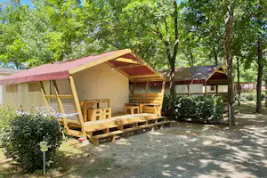 Campingunterkunft mit Veranda, Holzterrasse und Sitzbank im Schatten der Bäume, mit Zeltplane und kleinen Fenstern.
