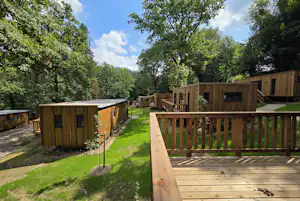 Holzhütten auf dem Campingplatz, Terrasse mit Holzgeländer, Rasenweg und Bäume im Hintergrund.