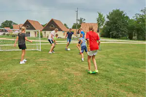Kinder und Erwachsene spielen Fußball auf dem Rasen vor Camping-Häusern, im Hintergrund Zaun und Bäume.