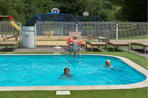 Kinder im blauen Schwimmbecken eines Campingplatzes, daneben Liegen auf Terrasse und Spielbereich mit Zaun und Rutsche.