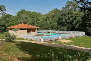 Schwimmbecken im Campingbereich, eingezäunt mit Metallgittern, daneben ein Gebäude mit Holzveranda und Ziegeldach.
