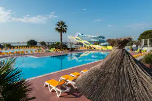 Schwimmbad mit türkisblauem Wasser, Sonnenliegen und gelben Rutschen, im Hintergrund Palmen und ein Wasserpark-Bereich.