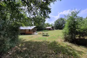 Zwei Zeltunterkünfte mit Vorzelten auf einer Wiese, Holztisch und Sitzbank, umgeben von Bäumen und Hecken.