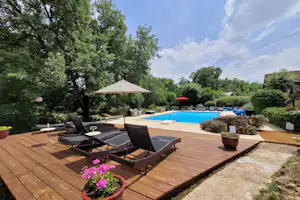 Poolbereich mit blauem Becken, Sonnenliegen und Sonnenschirm auf Holzterrasse, umgeben von Hecken und Bäumen.