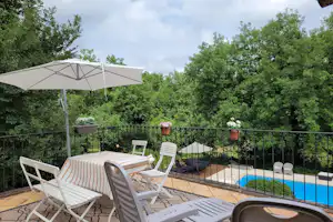 Terrasse mit Sonnenschirm, Liegestühlen und Tisch vor einem eingezäunten Pool, umgeben von Bäumen im Hintergrund.