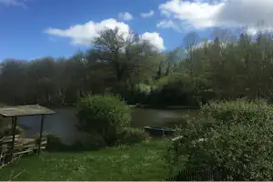 Blick auf einen Camping-See mit Ruderboot am Ufer, umgeben von Bäumen und einem Steg.
