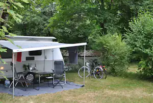 Wohnwagen mit Vorzelt und Tisch auf einer gepflasterten Fläche, daneben Fahrräder im Grünen unter Bäumen.