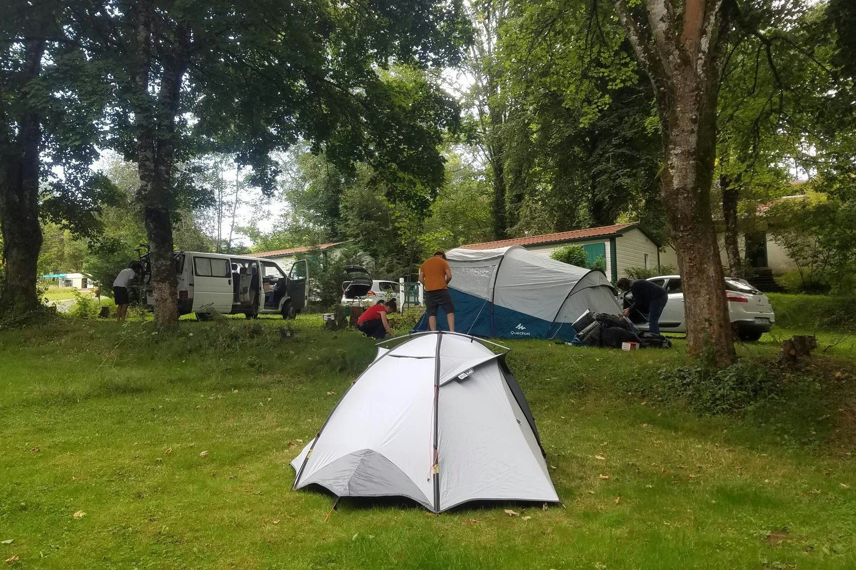 Campingzelt auf einer Wiese im Grünen, im Hintergrund Wohnwagen und Autos zwischen Bäumen.