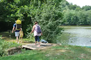 Zwei Wanderer gehen über einen kleinen Steg am See, im Hintergrund Bäume und grünes Ufer.