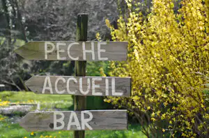 Holzwegweiser mit Aufschriften PÊCHE, ACCUEIL und BAR, im Garten mit gelben Blüten und Rasen.