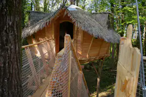 Holzhütte mit Strohdach und Holzbank am Eingang, umgeben von Bäumen, mit Netzgeländer und Holzstufen im Campingbereich.