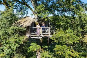 Baumhaus auf Stelzen im Grünen mit Strohdach und Balkon, davor Geländer und Treppe im Campingbereich.