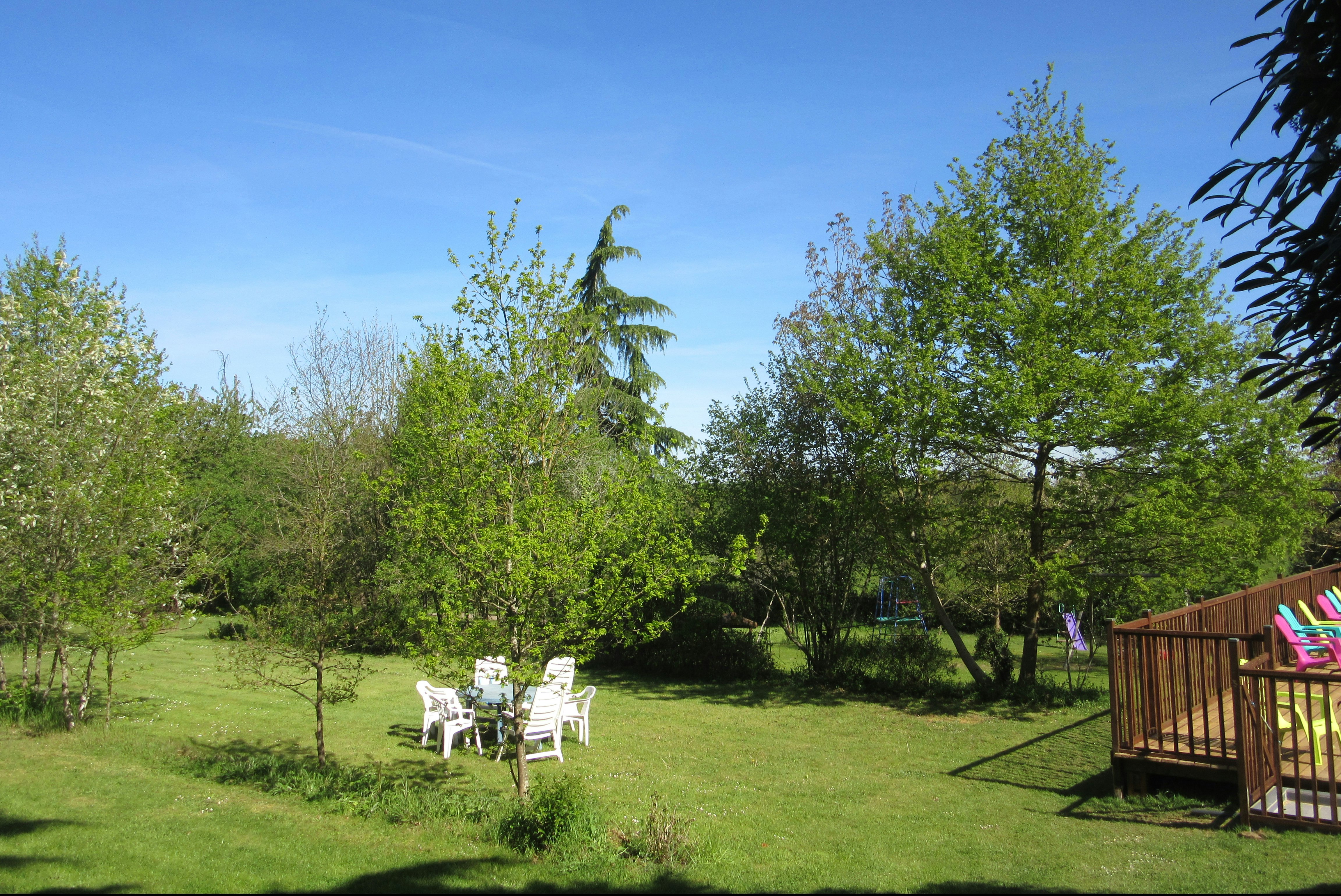 Grüner Campinggarten mit Sitzgruppe aus weißen Stühlen und Blick auf eine Holzterrasse, umgeben von Bäumen und Rasen.