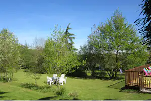 Grüner Campinggarten mit Sitzgruppe aus weißen Stühlen und Blick auf eine Holzterrasse, umgeben von Bäumen und Rasen.