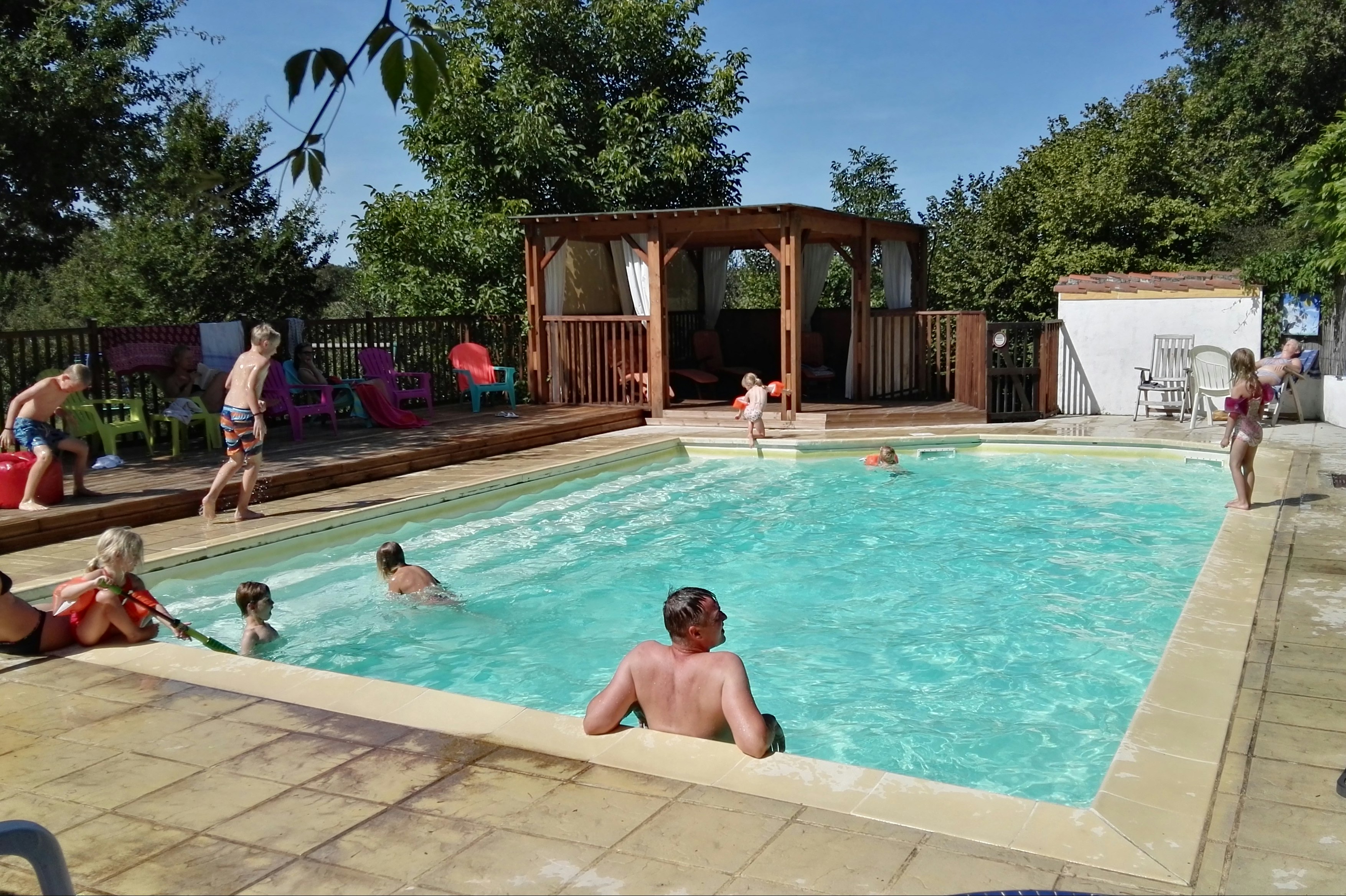 Schwimmbecken auf dem Campingplatz mit Holzpavillon, Liegestühlen und bunten Gartenstühlen im Hintergrund.