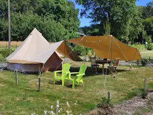 Zelte auf einem Campingplatz im Grünen, mit Tisch und grünen Stühlen, umgeben von Bäumen und einem eingezäunten Bereich.