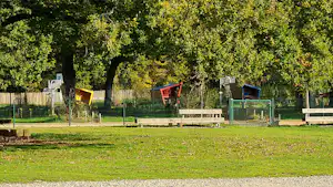 Grüne Rasenfläche mit Holzbalken und Sitzgelegenheiten am Campingplatz, dahinter ein Zaun und Bäume.
