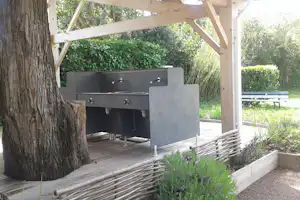 Gasgrill unter Holzüberdachung auf einer Terrasse, mit Sicht auf Hecken und Gartenbank im Hintergrund.