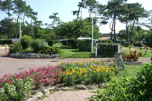 Blumenbeete und Rasenfläche im Campinggarten, gepflasterter Weg, Hecken und hohe Bäume mit Laternen.