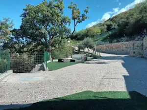 Campingplatz mit Kiesweg, Rasenfläche und Zaun, daneben Bäume und ein Steinmauerhang mit Blick in die Landschaft.