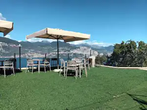 Terrasse mit Rasen, mehreren Stühlen und Sonnenschirmen, Blick auf Berge und einen Zaun am Campingplatz.