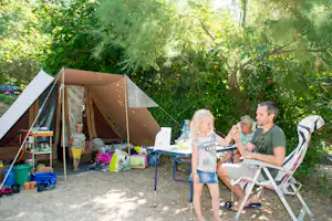 Campingzelt im Grünen, Familie sitzt am Tisch mit Getränken, Sonnenschirm und Liegestuhl neben dem Zelt.