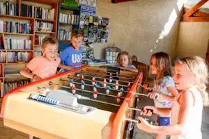 Kinder spielen am Tischkicker in einer Camping-Ecke, im Hintergrund Bücherregal und Infowand mit Flyern.