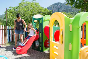 Kinder spielen an einer bunten Rutsche auf dem Campingplatz, im Hintergrund Bäume und Berge.