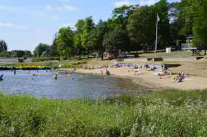 Sandstrand am See mit vielen Badegästen, Liegetüchern und Sonnenschirmen, im Hintergrund Bäume und ein Steg.