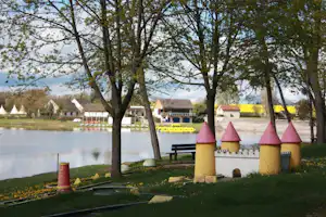 Blick auf den See mit Bäumen und bunten Spieltürmen am Ufer, im Hintergrund Zelte und Gebäude.