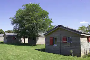 Holz-Campinghaus mit roter Tür und Fensterläden, Rasenfläche davor und Baum im Hintergrund.