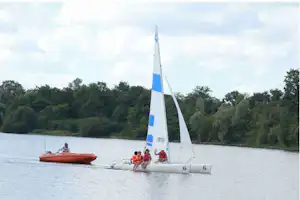 Ein Segelboot auf einem See, daneben ein Ruderboot; im Hintergrund Wald und ruhiges Wasser.