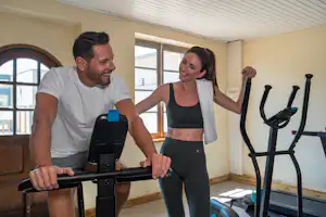 Zwei Personen trainieren an einem Fitnessgerät in einem hellen Camping-Fitnessraum mit großen Fenstern und Sportmatten.
