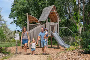 Familie am Spielplatz mit Holzspielhaus und Rutsche auf dem Campingplatz, umgeben von Bäumen und Wegen.
