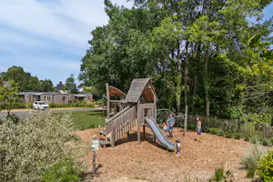 Spielplatz mit Holzspielturm und Rutsche auf einem Campingplatz, umgeben von Bäumen, Wegen und einem kleinen Gartenbereich.