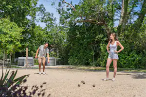 Zwei Personen spielen Boule auf einem Kiesplatz im Grünen, mit Bäumen und Hecken im Hintergrund.
