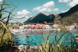 Badebereich am Campingplatz mit Schwimmern im See, Liegeflächen und Gebäuden am Ufer, im Hintergrund Berge und Wald.
