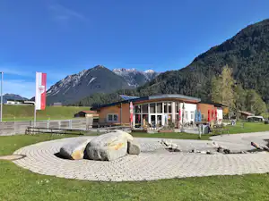 Rund gepflasterter Feuerstellenbereich mit großen Steinen vor einem Campinggebäude, Wiese, Zäunen und Bergen im Hintergrund.