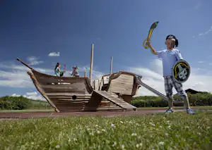 Kinder auf einem hölzernen Spielboot, ein Junge mit Piratenhut und Schwert, andere Kinder klettern im Hintergrund.