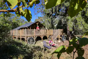 Holzhütte auf Stelzen mit Veranda, Leiter und Wagenrad, im Grünen zwischen Bäumen; im Vordergrund Stühle und Tisch.