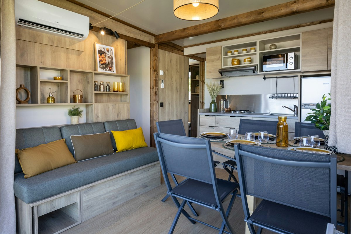 Wohnbereich mit grauem Sofa, gelben Kissen und Esstisch im hellen Holzbungalow, Küche mit Schränken und Pflanzen im Hintergrund.