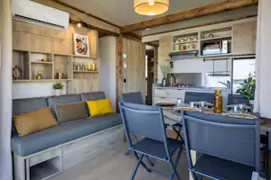 Wohnbereich mit grauem Sofa, gelben Kissen und Esstisch im hellen Holzbungalow, Küche mit Schränken und Pflanzen im Hintergrund.