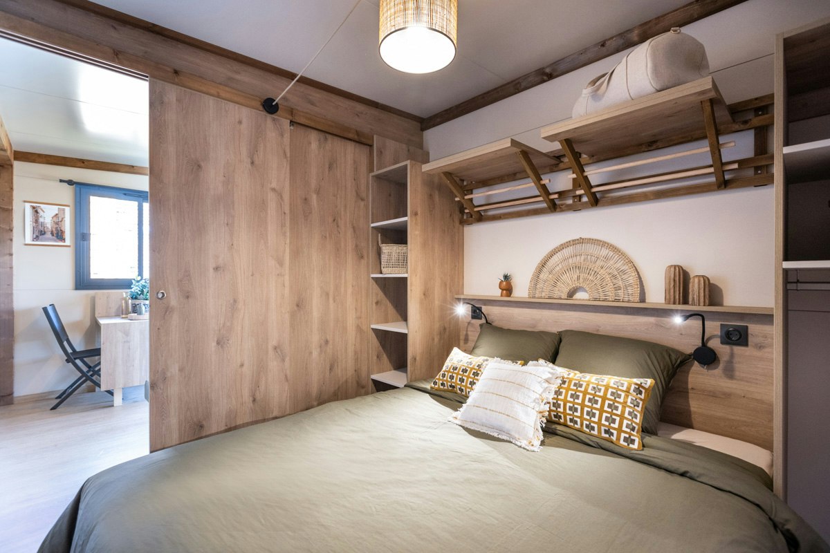 Gemütliches Schlafzimmer mit Doppelbett, Holzverkleidung und Regalen, darüber eine Lampe; im Hintergrund ein Fenster und ein kleiner Tisch.