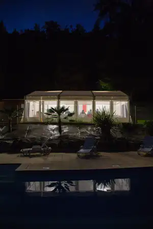 Beleuchtete Terrasse am Campingplatz mit Pool und Liegestühlen vor einem Wohnwagen-Gebäude bei Nacht.