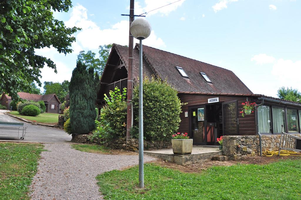 Campinggebäude mit Eingang, Holzfassade und Schild, davor Sitzbank und Steintreppe, umgeben von Rasen und Büschen.