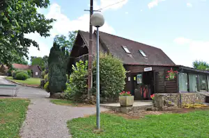 Campinggebäude mit Eingang, Holzfassade und Schild, davor Sitzbank und Steintreppe, umgeben von Rasen und Büschen.