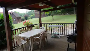 Überdachte Camping-Terrasse mit Tisch und Stühlen, Blick auf Rasen, Bäume und kleine Gebäude im Hintergrund.